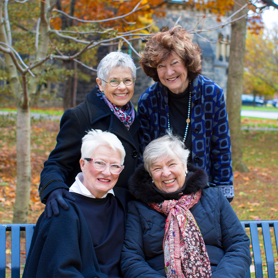 Esther Atkin, Lynn Corbey, Karen Jones and Aita Moore smile as they pose on a bench on Front Campus near autumn trees