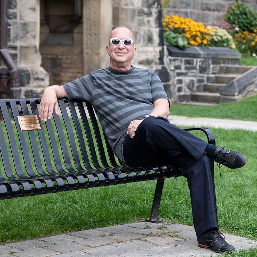 Paul Shaffer smiles as he sits on a bench outside University College, with one arm stretched out along the top